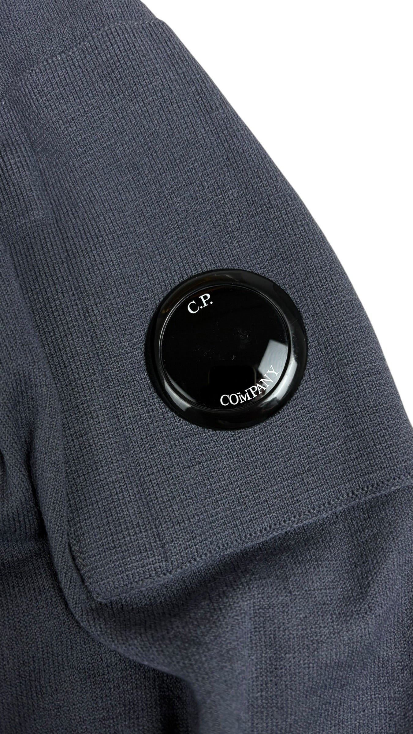 Gros plan sur l'ecusson lentille noir C.P Company cousu sur la manche du sweat, maille cotelee bleu marine, vue detail