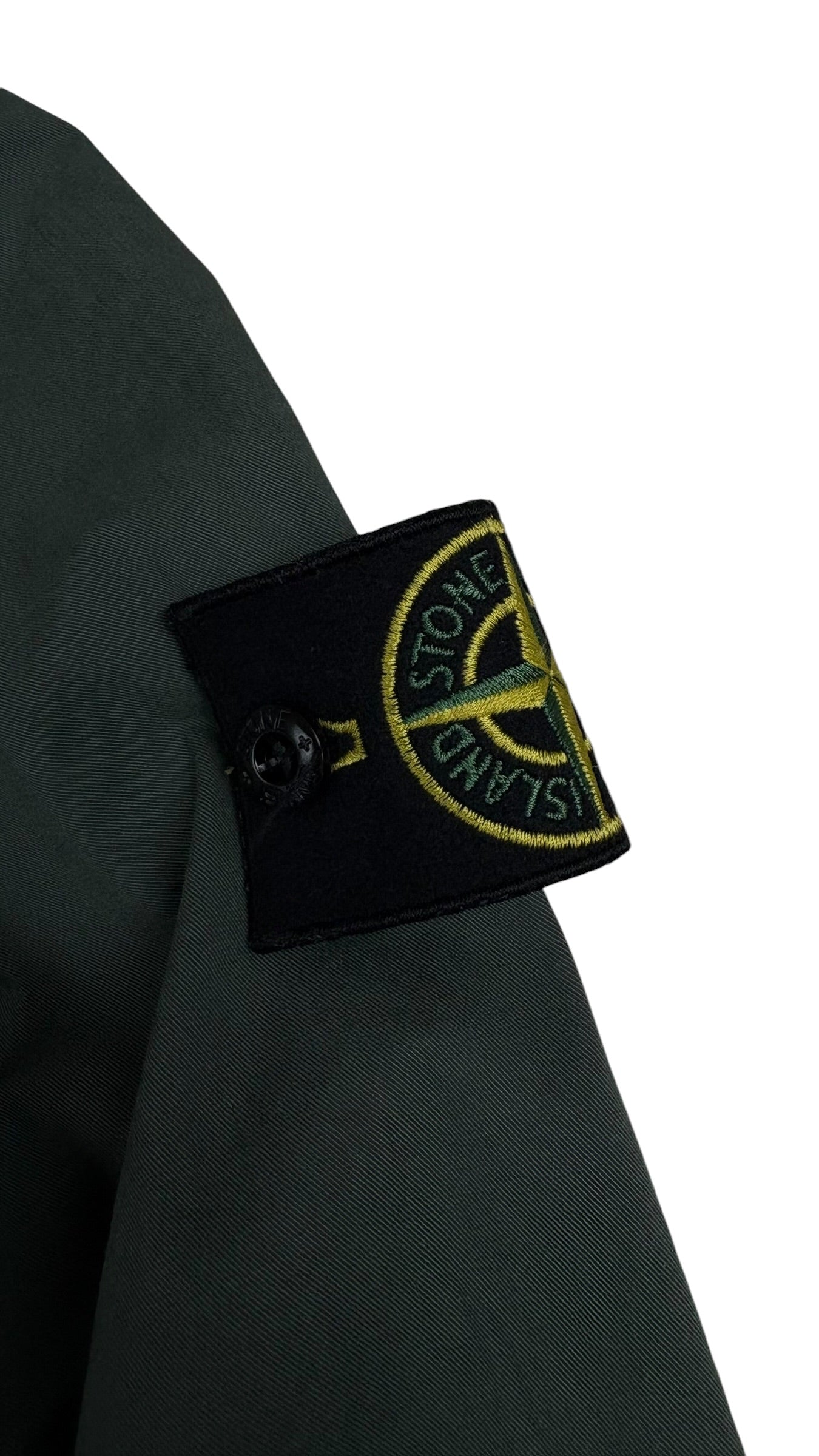 Gros plan sur l'écusson boussole Stone Island jaune et vert, badge amovible fixé par boutons sur la manche de la surchemise kaki, vue détail