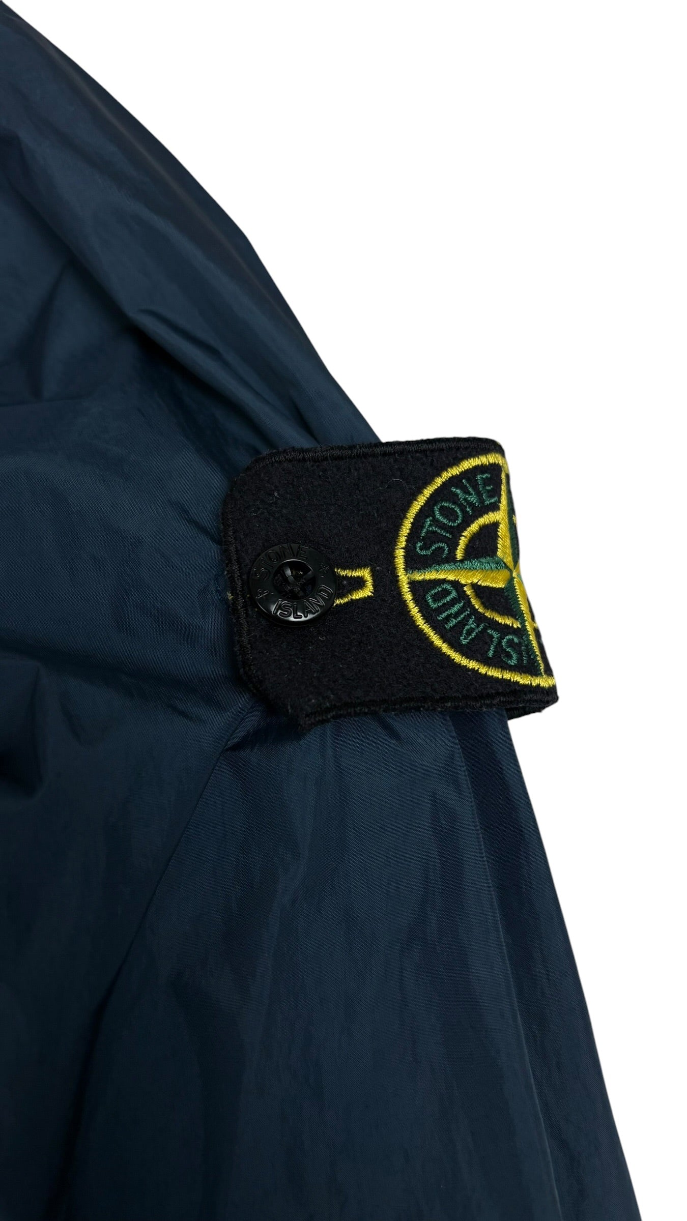 Gros plan sur l'écusson boussole Stone Island jaune et vert, badge amovible fixé par boutons sur la manche de la veste coupe-vent bleu marine, vue détail