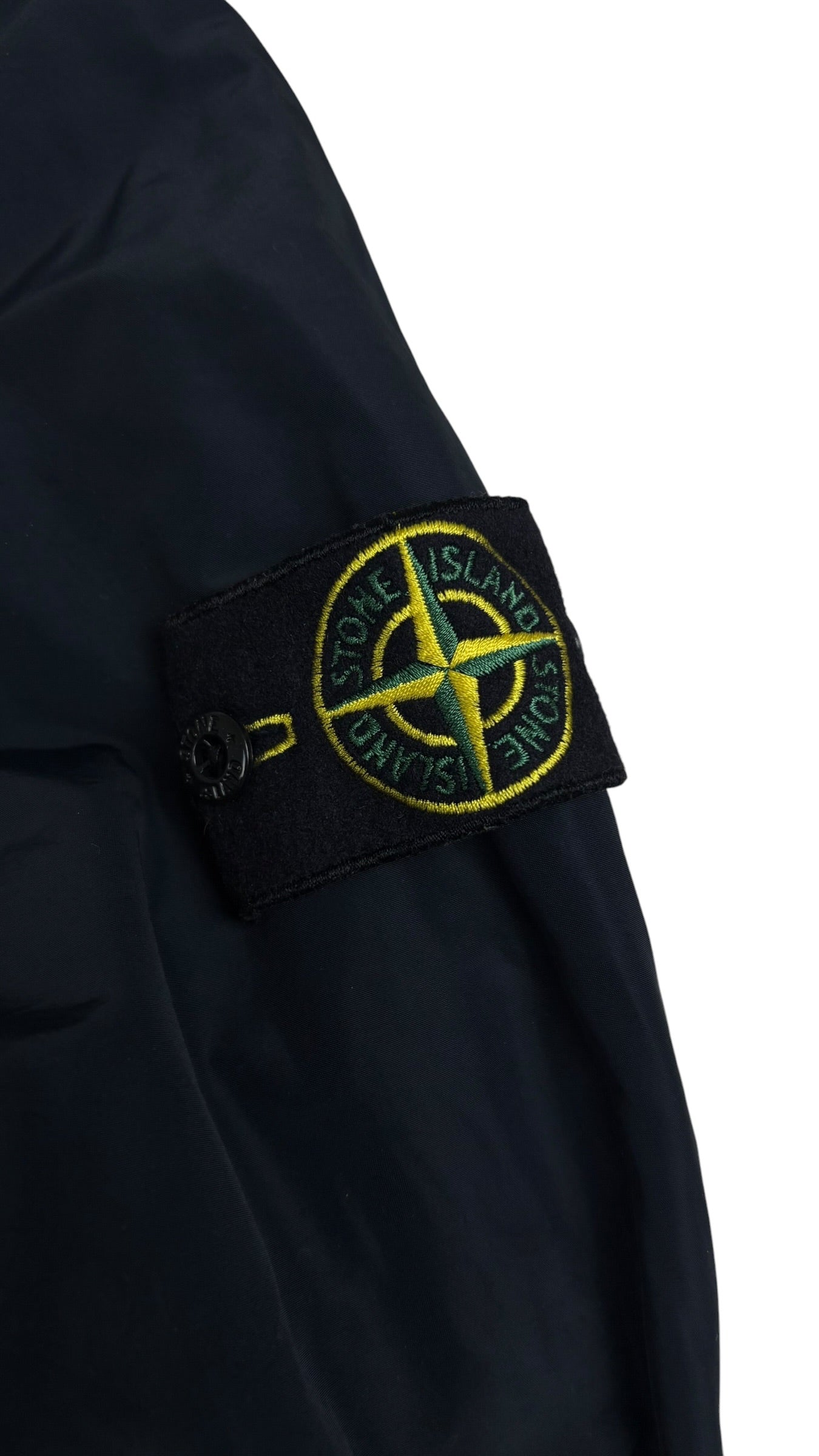 Gros plan sur l'écusson boussole Stone Island jaune et vert, badge amovible fixé par boutons sur la manche de la veste bleu marine, vue détail